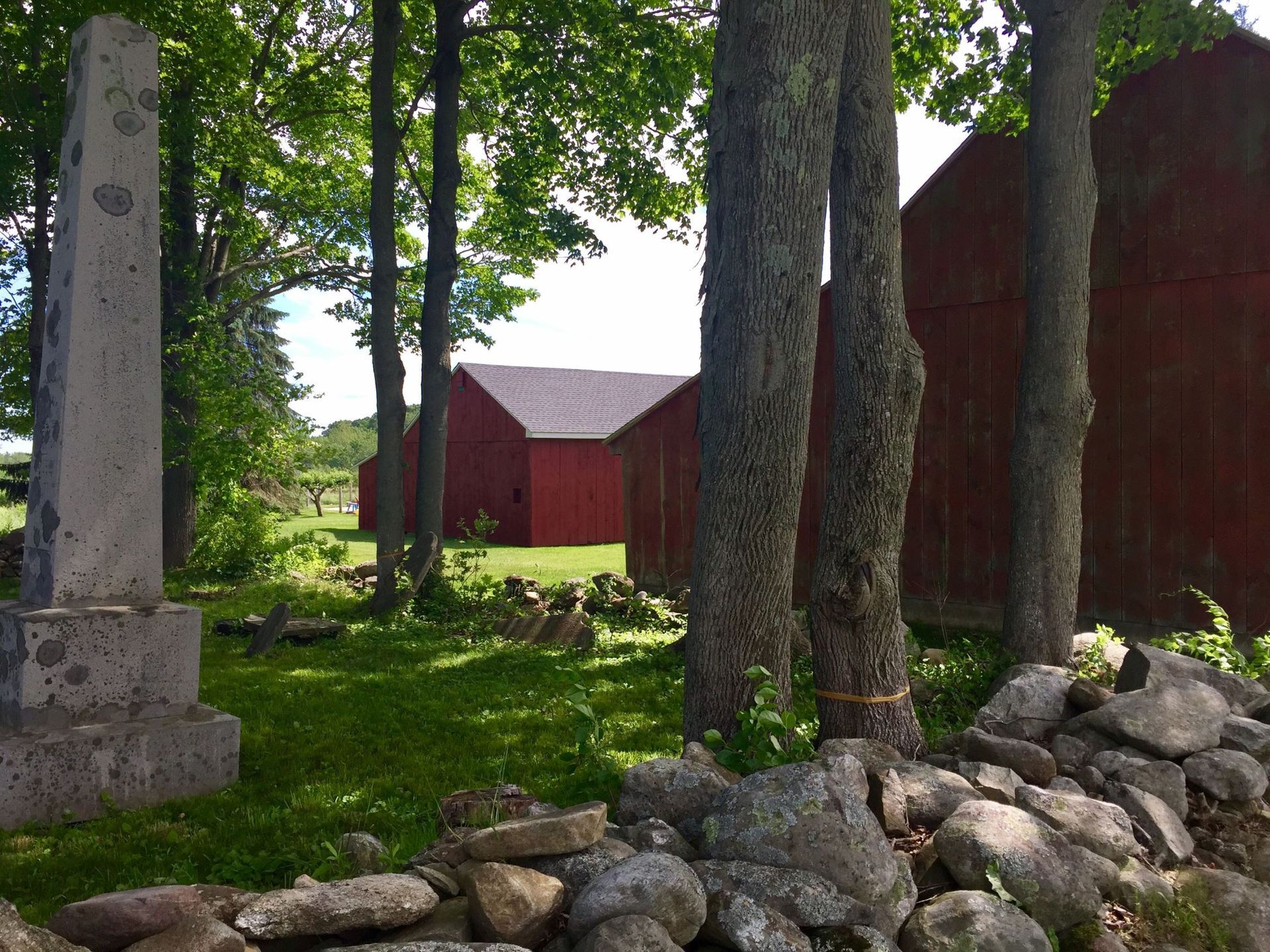 A stone obelisk is surrounded by trees and rocks in front of a red barn