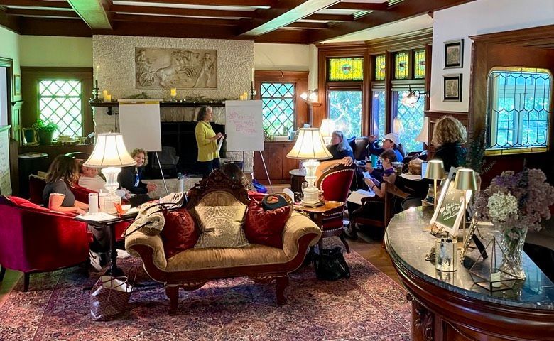 Group meeting in a vintage living room. People seated, one woman presents. Fireplace, stained glass windows.