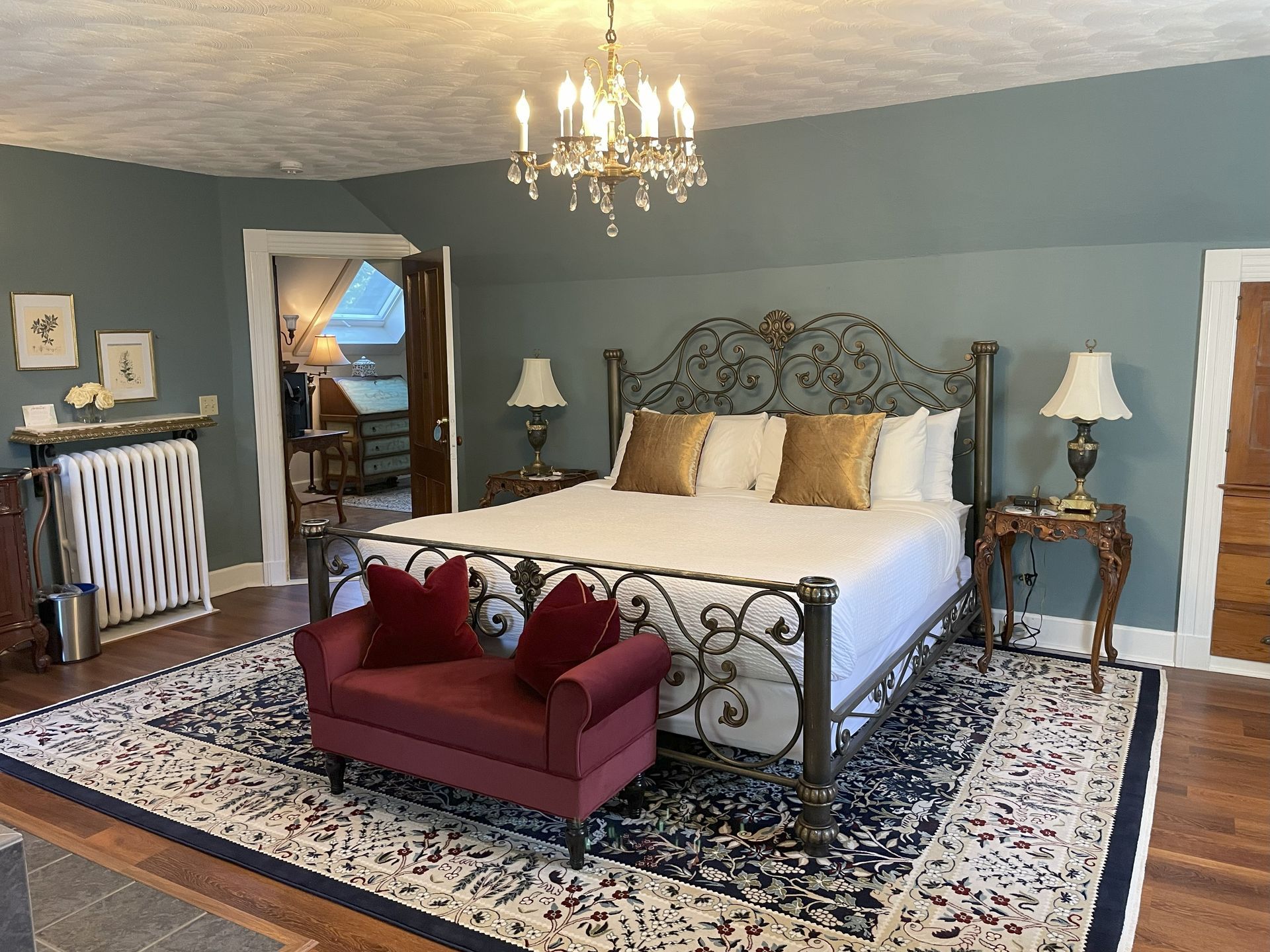 Elegant Victorian bedroom with a wrought iron bed, teal walls, a chandelier, and a red bench.