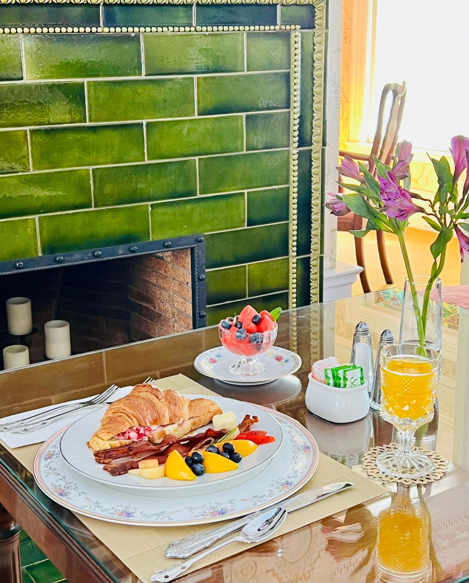 Breakfast on a table with a croissant, fruit, and juice near a green tiled fireplace and flowers.