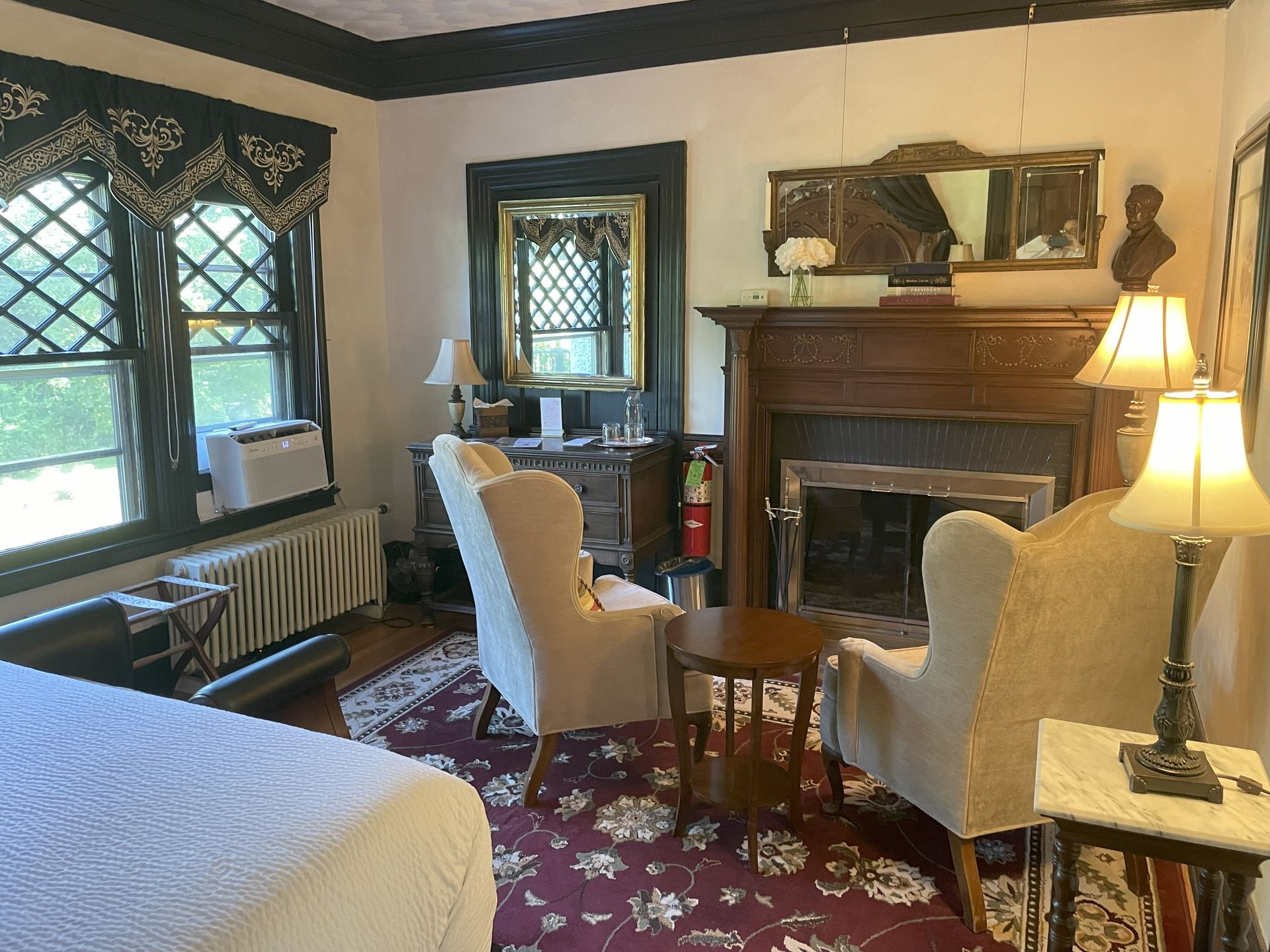 Cozy hotel room with fireplace, chairs, and window. Dark trim, floral rug, and lamps.