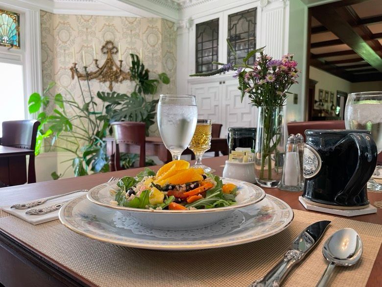 Salad with orange slices, water glass, flowers, and coffee cup on a dining table in a sunlit room.