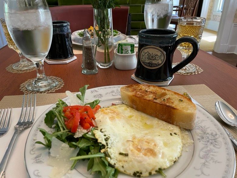 Breakfast plate with fried eggs, greens, toasted bagette, and coffee mug on a table setting.