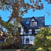 A large white house with a black roof is surrounded by trees on a sunny day.