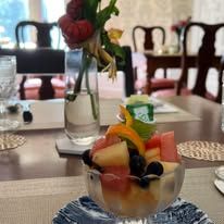 A crystal bowl with fruit salad is sitting on a plate on a table with fresh flowers in the background.