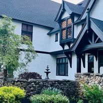 A large white house with a black roof and a stone wall in front of it.