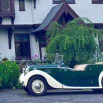 A green and white vintage car is parked in front of a Tudor-style white mansion.
