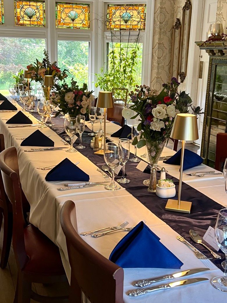 Long table set for a formal dinner, with flowers, navy napkins, gold lamps, and ornate stained glass windows.