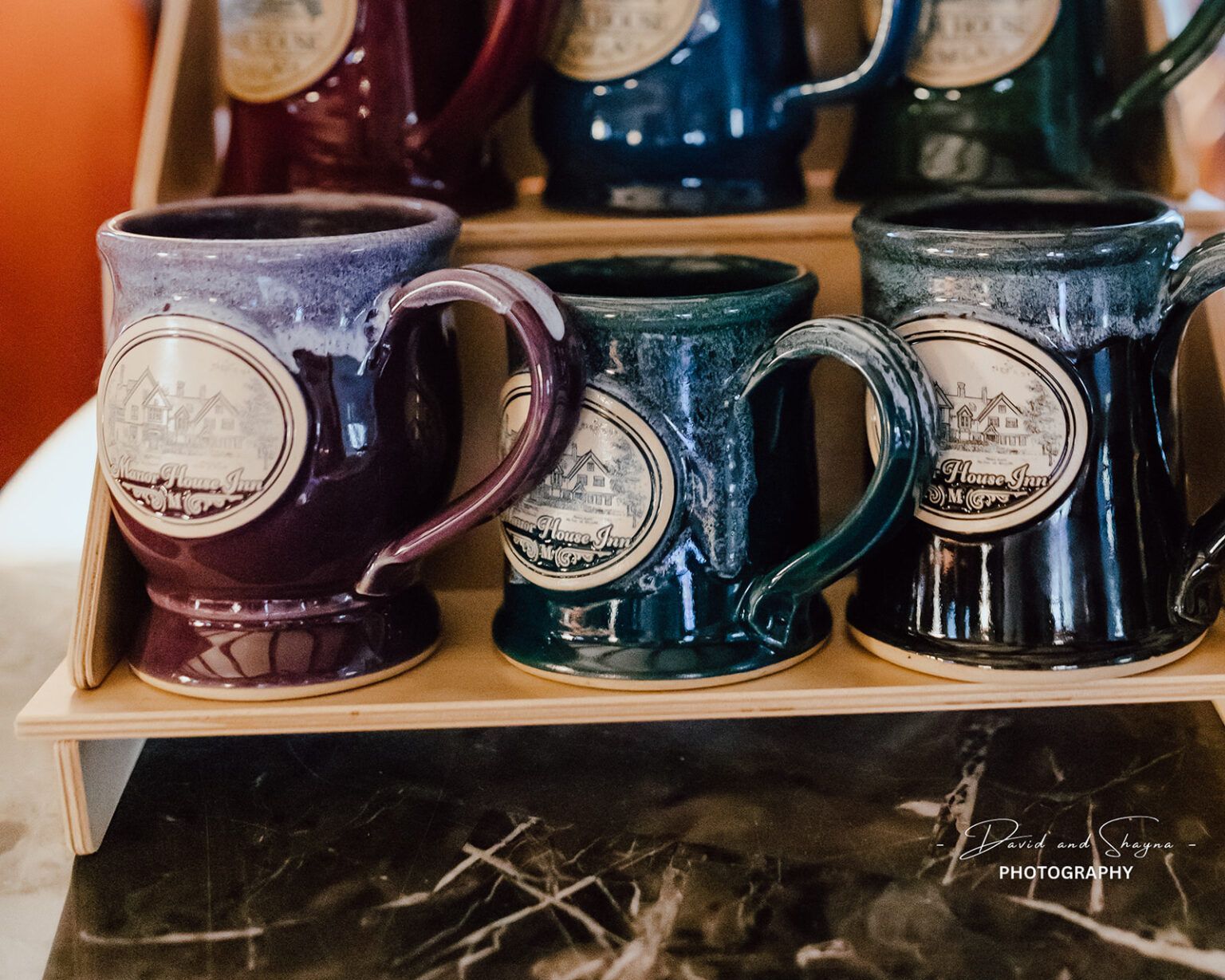 A row of coffee mugs with Manor House Inn logos sitting on top of a wooden shelf.