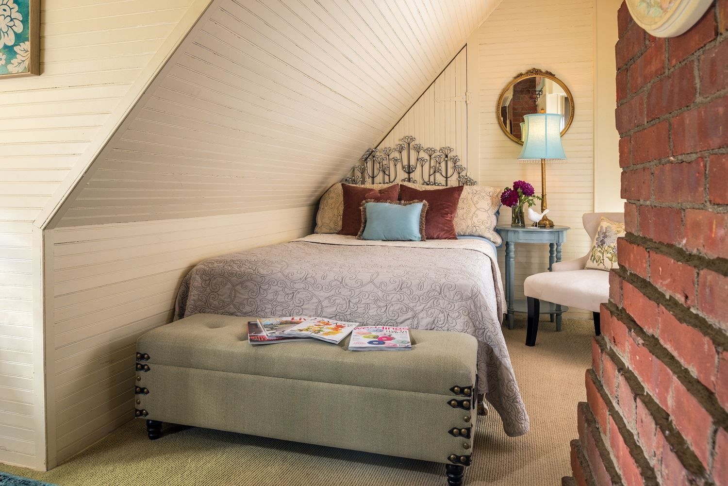 A bedroom with a bed , ottoman , chair and brick wall.