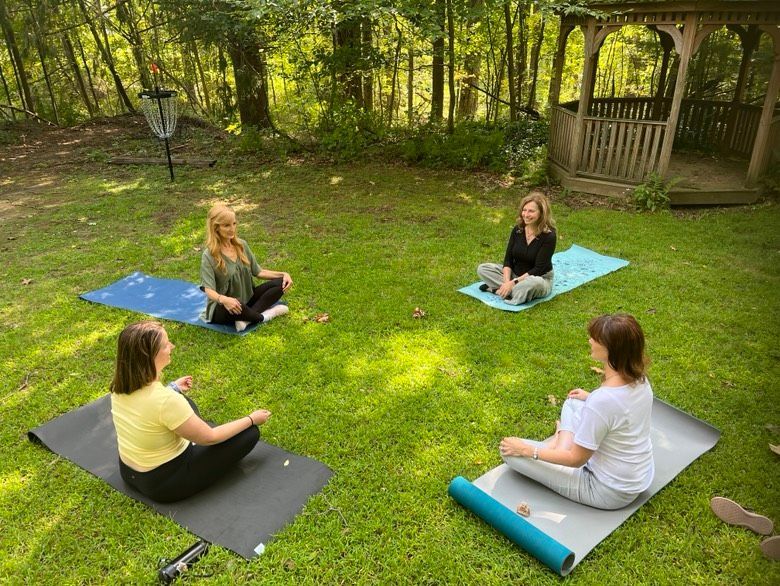Four women meditate outdoors on mats in a grassy yard, near a gazebo, surrounded by trees.