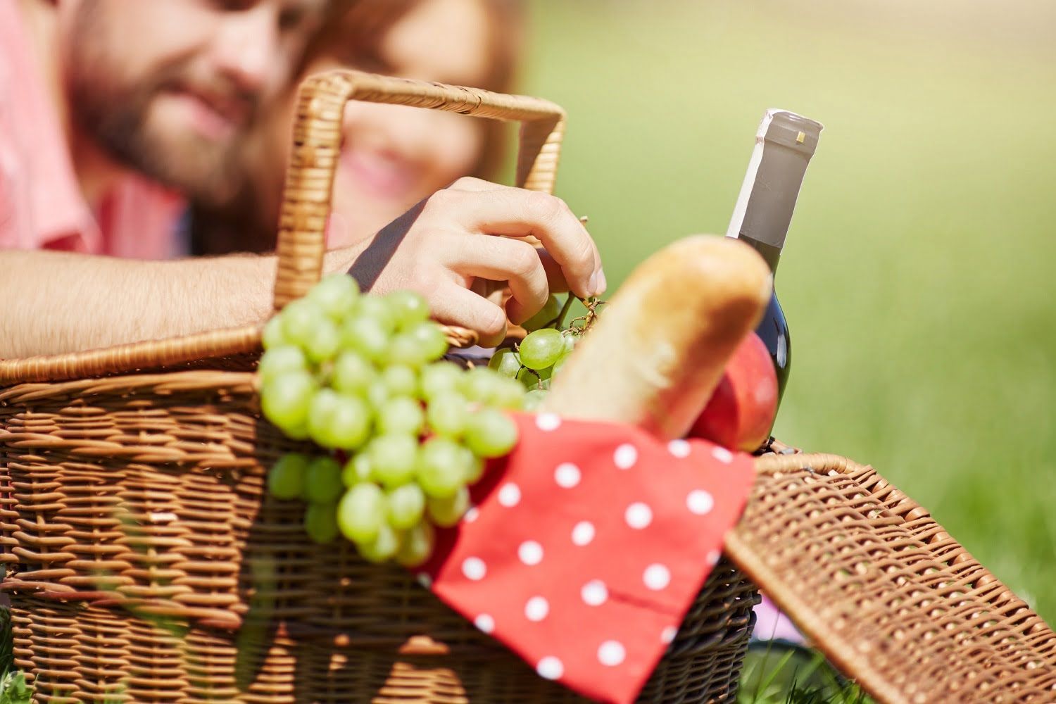 A man is holding a picnic basket with grapes , bread and a bottle of wine.