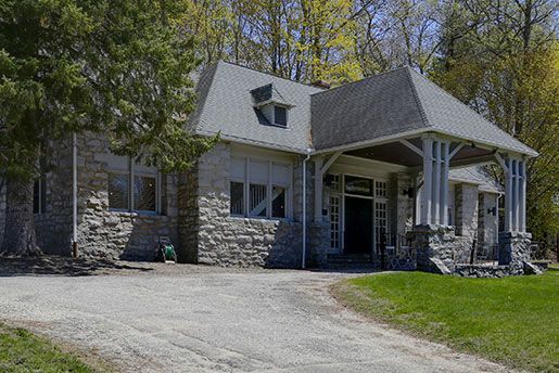 A large stone house with a driveway leading to it is surrounded by trees.