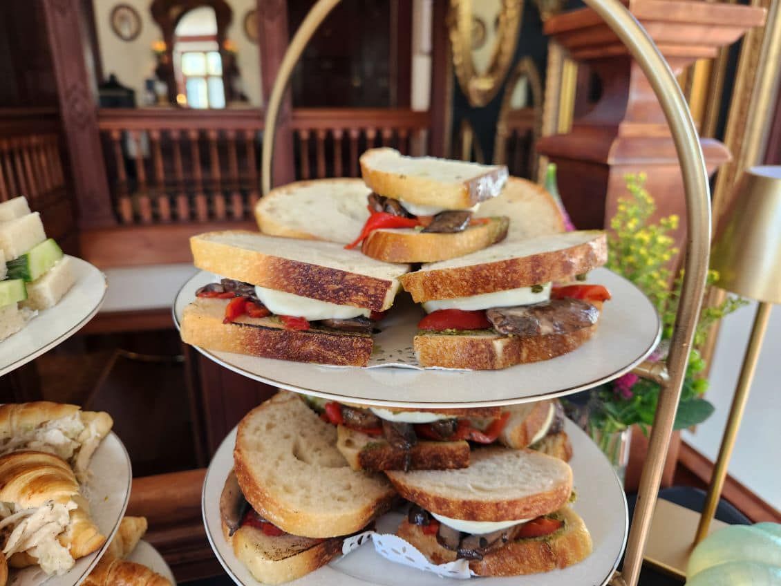 Tiered tray of sandwiches: bread, mozzarella, roasted red pepper, and eggplant. Indoors, ornate setting.