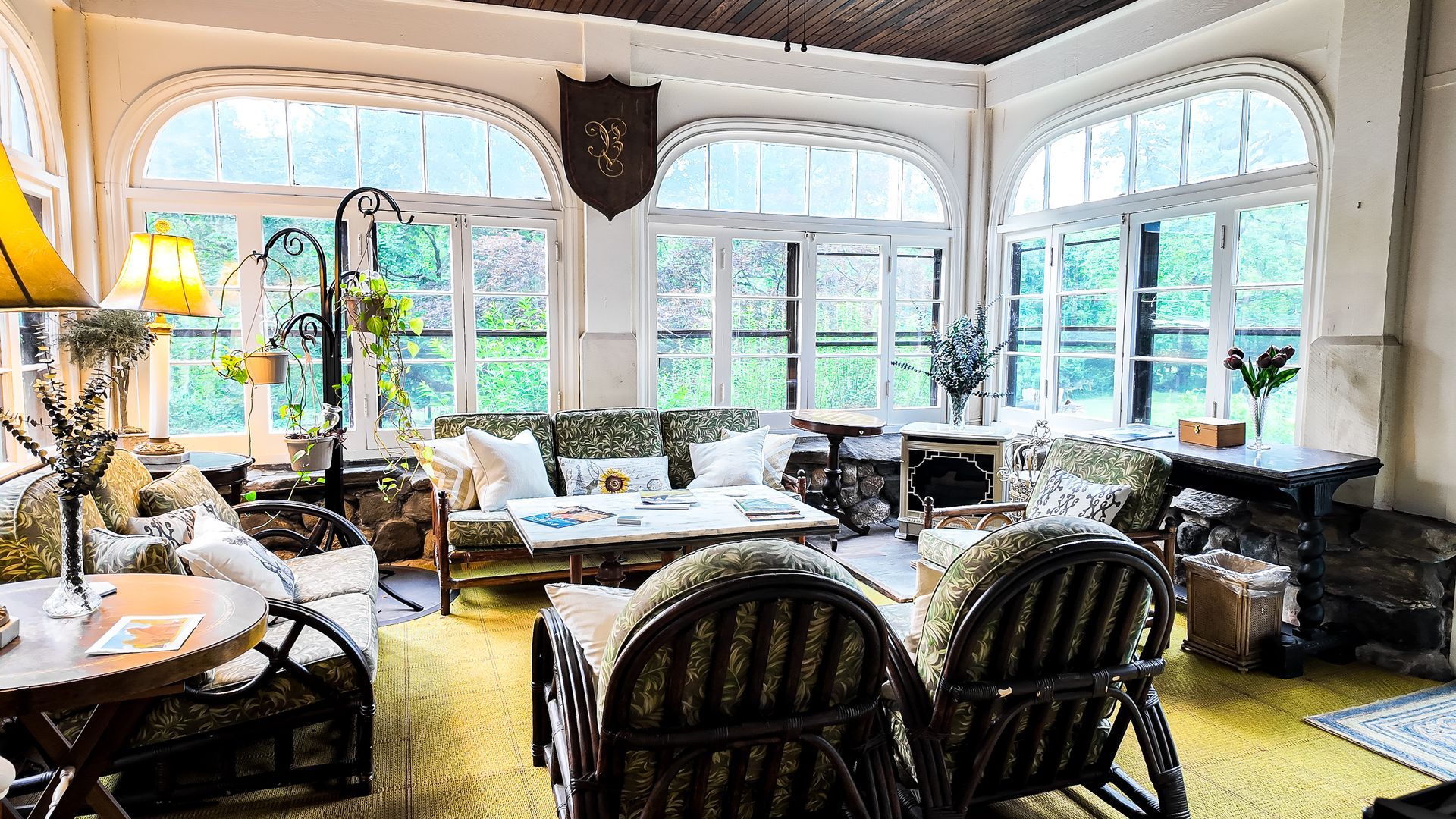Sunroom with wicker furniture, patterned cushions, large windows, and a shield emblem.