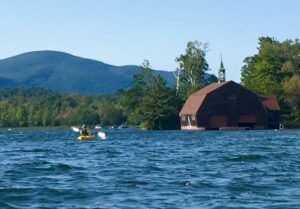 A person is paddling a kayak on a lake with mountains in the background.