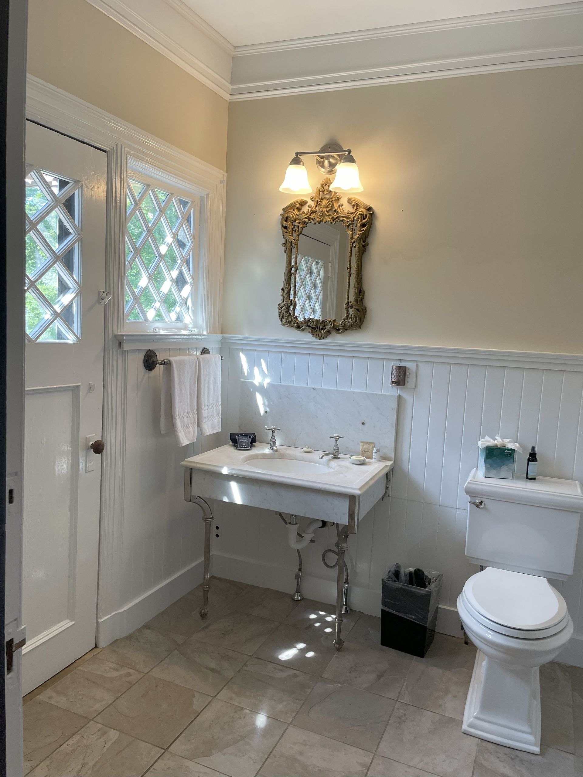 Bathroom with white fixtures, gold mirror, window, and marble-topped sink.