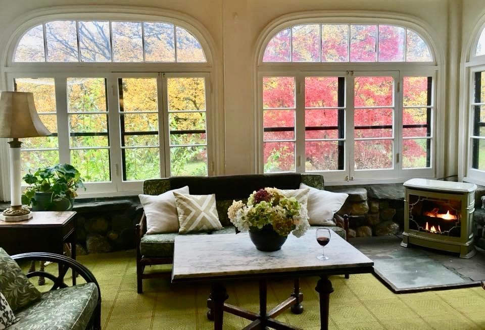 A sunroom with a couch, table, fireplace, and windows overlooking New England fall foliage.