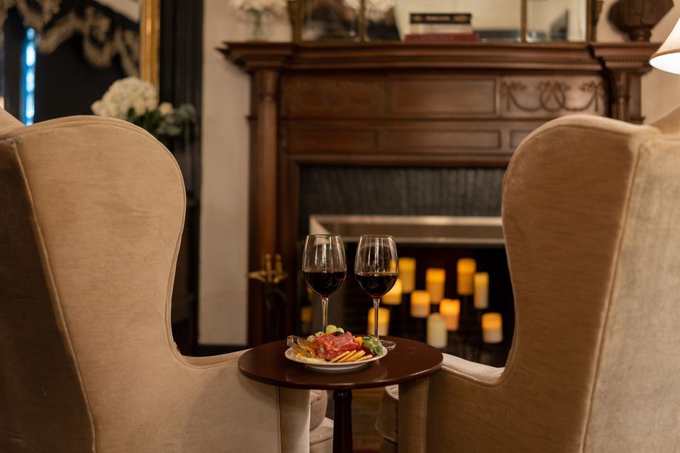 Two chairs sit in front of a fireplace, accompanied by two glasses of wine on a table.