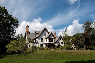 A large white Tudor-style house is sitting on top of a grassy hill surrounded by trees.