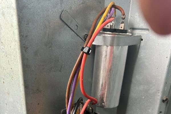 A cylindrical metal run capacitor installed inside a gray metal cabinet, connected by several colored electrical wires.