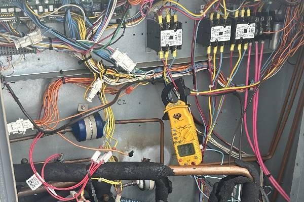 A yellow multimeter clamped onto a wire inside an open HVAC electrical control panel with various colored cables.