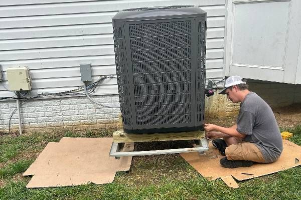 A technician in a cap kneels on cardboard, working on the base of an outdoor HVAC unit against a white siding house.