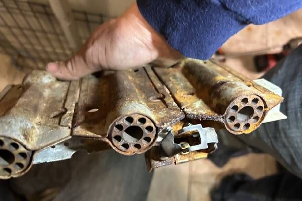 A hand holds a rusted, multi-tubular metal furnace burner assembly.