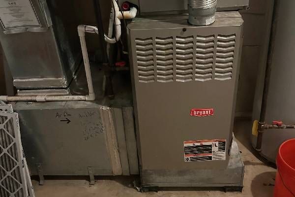A beige Bryant furnace unit stands next to a metal air duct and a white PVC drain pipe in a basement setting.
