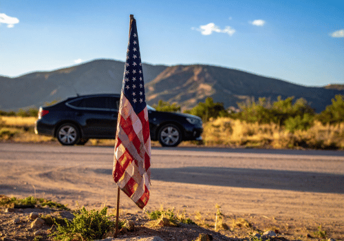 American flag beside a roadside with a parked SUV and mountains in the background.