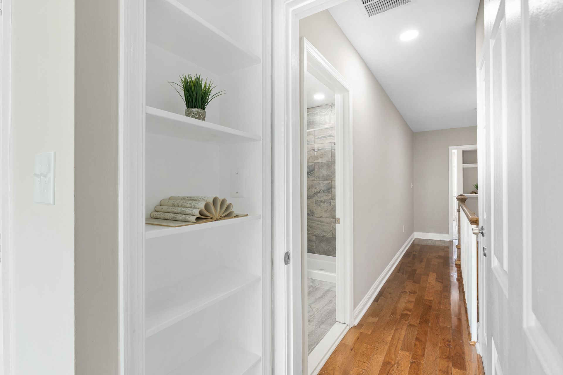 A hallway with hardwood floors and white shelves leading to a bathroom.