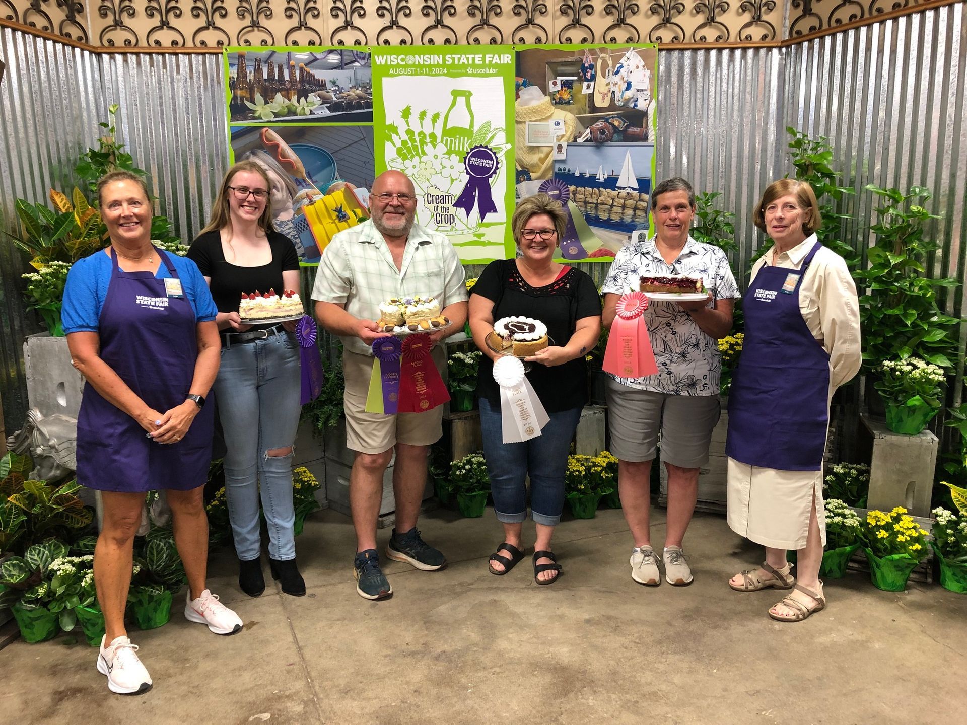 Six people hold baked goods at a fair. Green backdrop, flowers, and a banner.
