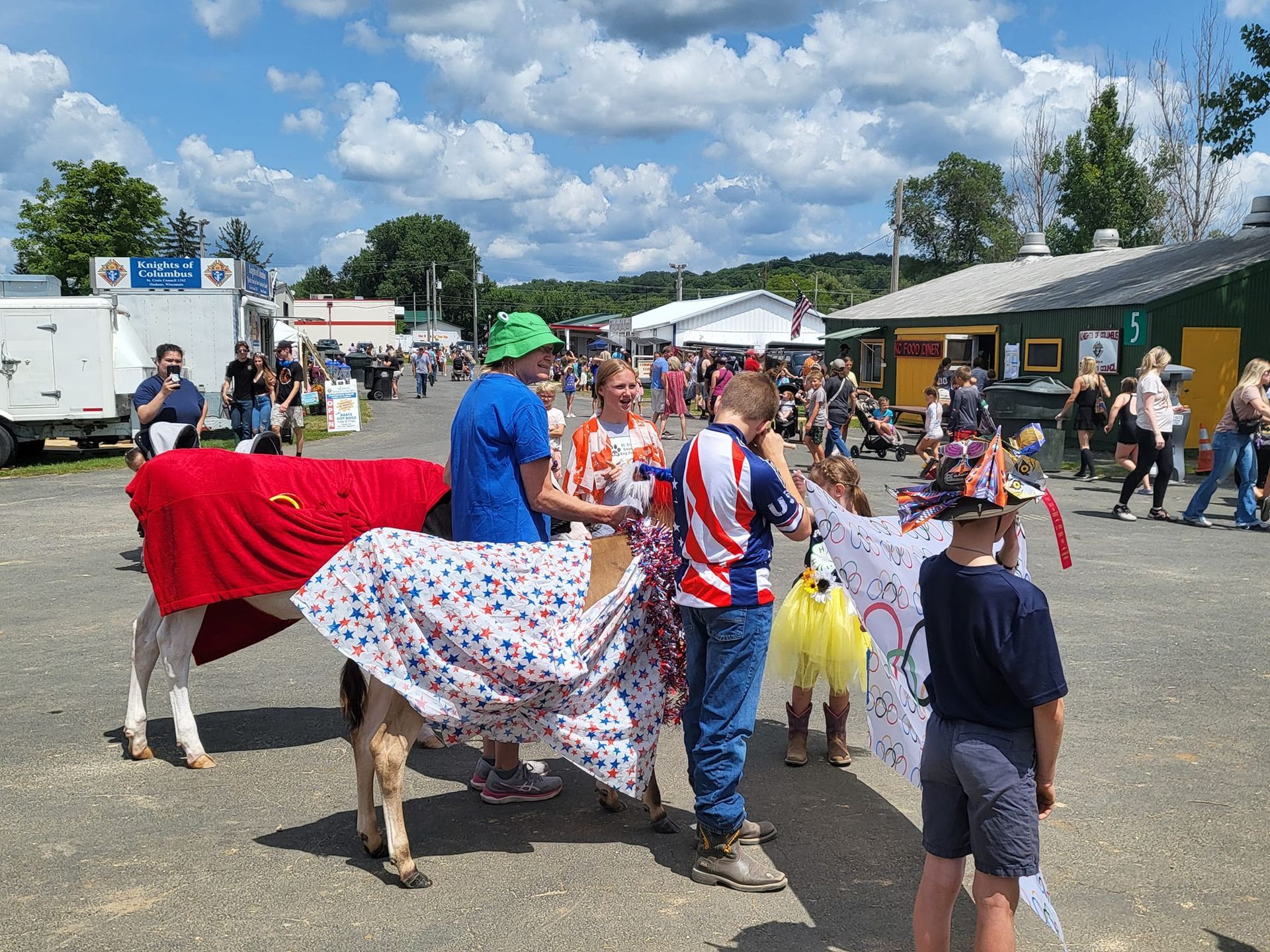 Children drape decorated blankets over a goat at an outdoor fair, people and vendor booths in background.