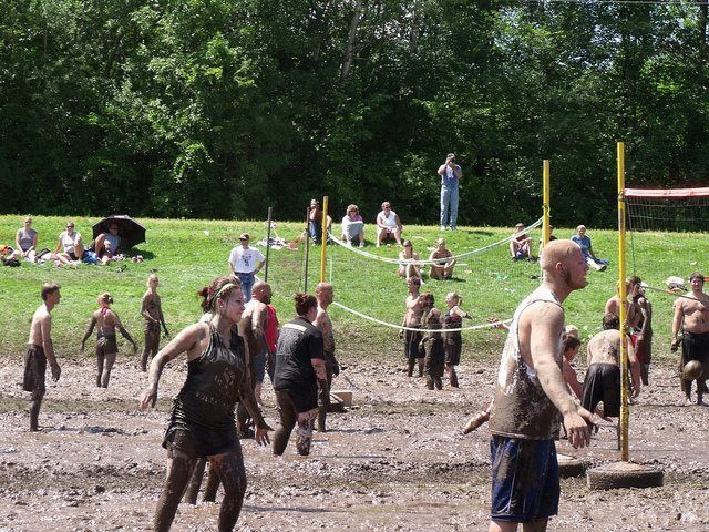 People playing mud volleyball on a grassy field with spectators and trees in the background.