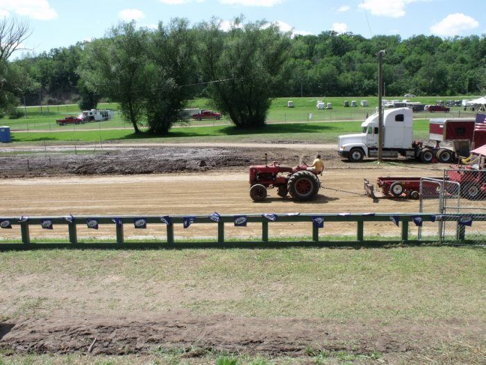 Red tractor pulling a weight sled at a tractor pull event.  A semi-truck and trees are in the background.