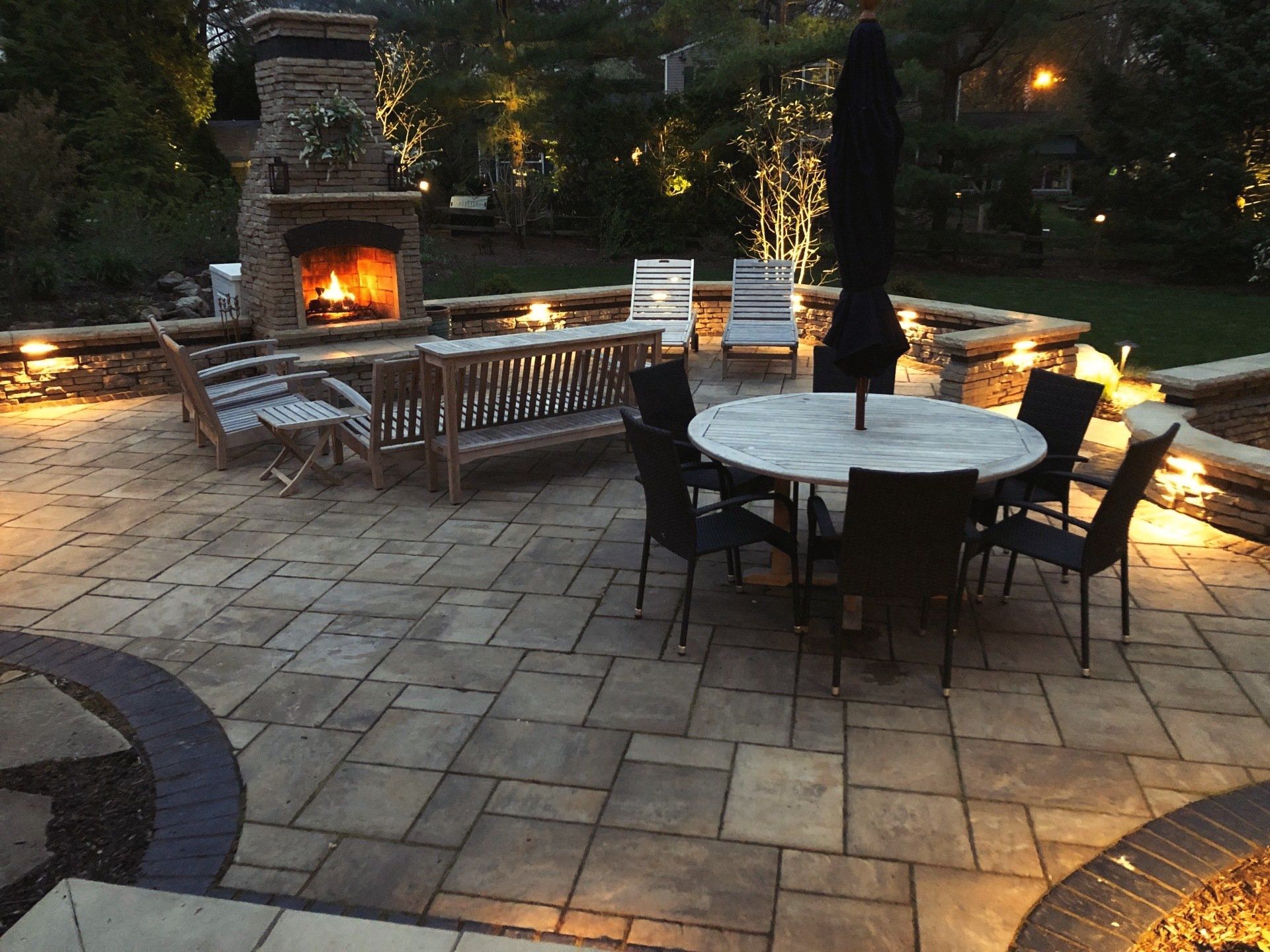A nighttime patio with a lit fireplace, seating area, and a round table with chairs. Warm lighting illuminates the space.