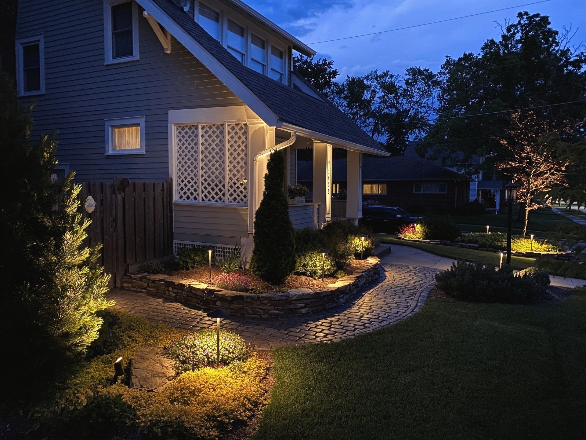 Lit-up house exterior at dusk, showcasing landscape lighting on bushes, pathway, and tree, creating a warm ambiance.
