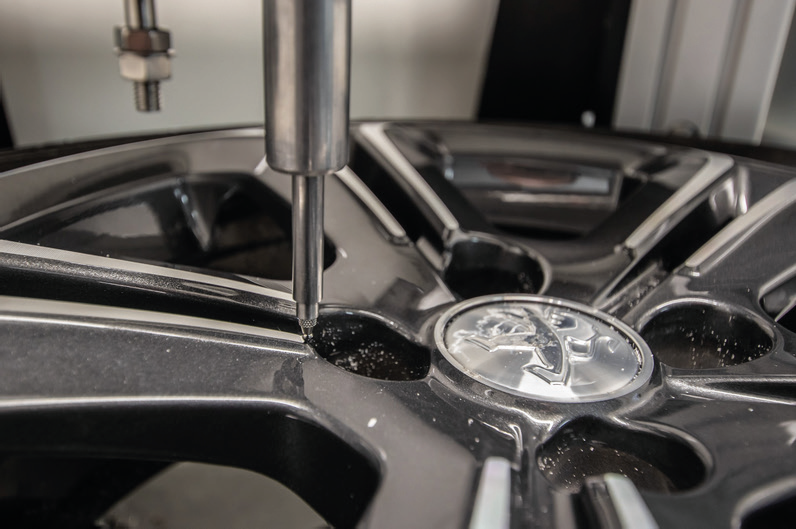 A machine tool drilling into a gray car wheel near the Peugeot logo.