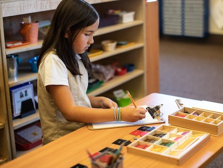 A young girl is sitting at a table writing in a notebook with montessori materials