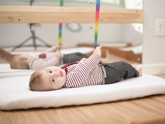 Montessori infant lying on a white mat, looking at the camera, with a mirror in the background reflecting the same scene.