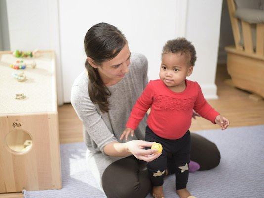 Montessori guide kneeling, helping toddler walk, holding a yellow toy, in a room with wooden furniture.