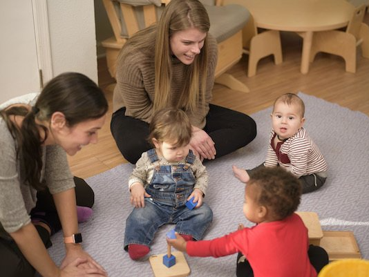 Two Montessori guides and three toddlers on a blue rug, playing. One child holds a toy.