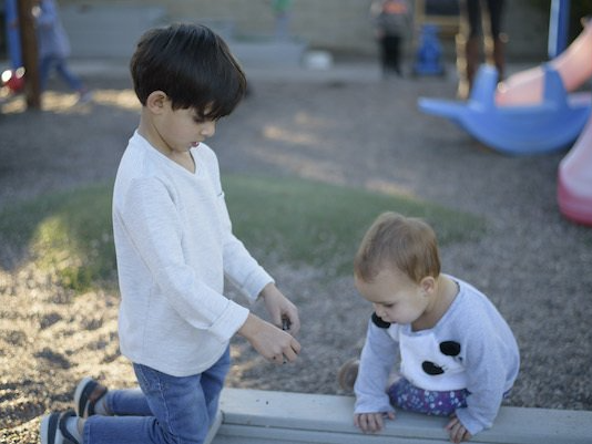 Montessori child kneeling, pointing at something for baby sitting on the ground at a playground.