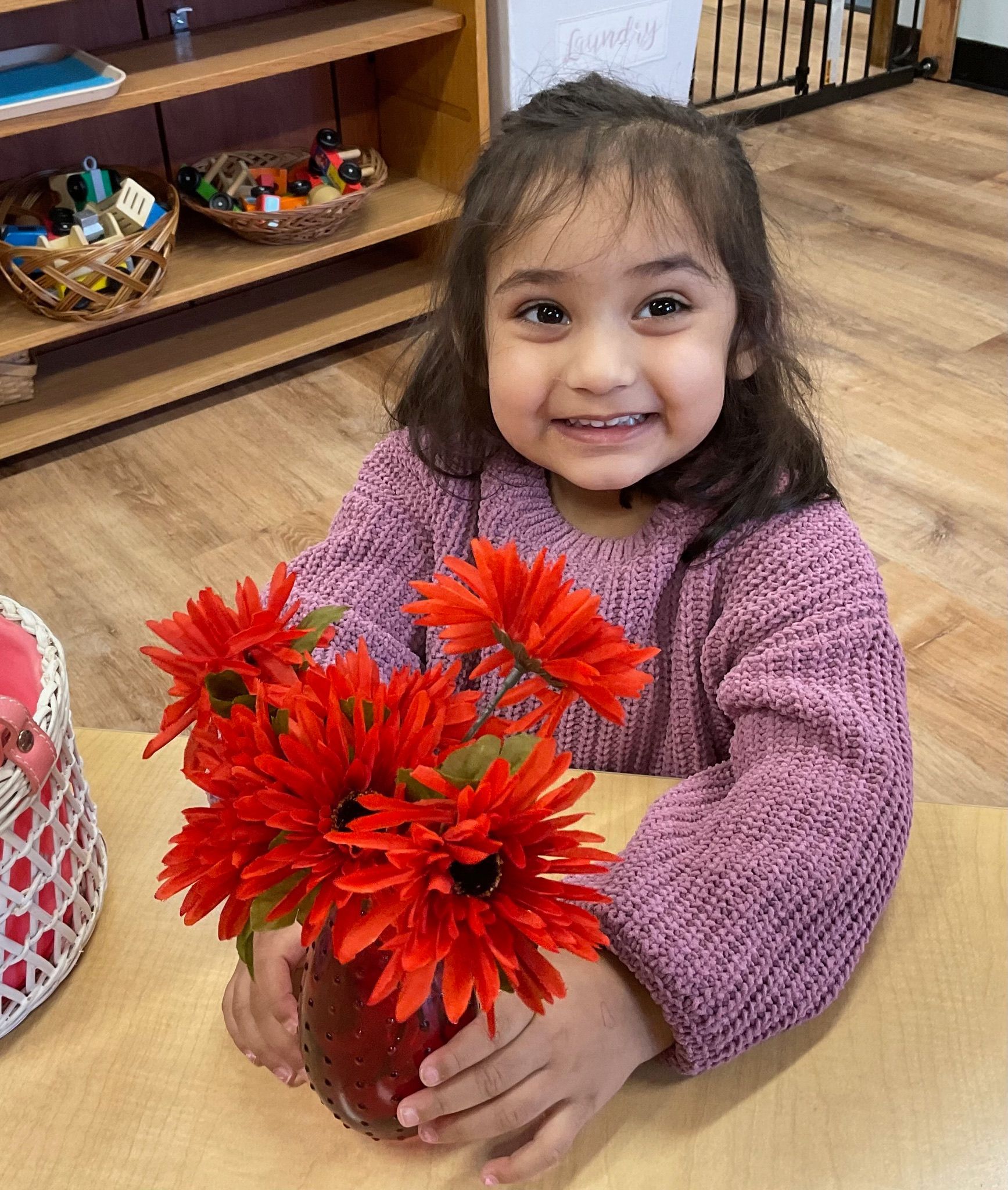 Montessori child is sitting at a table holding a vase of red flowers.