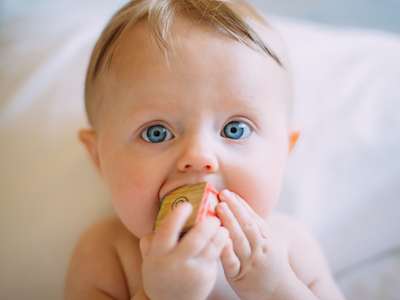Baby with bright blue eyes, holding a wooden block in their mouth.