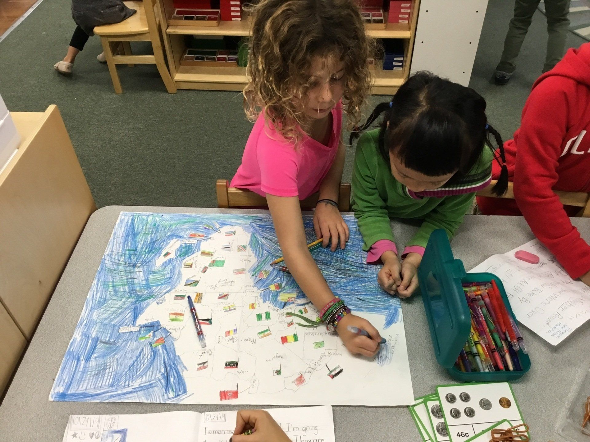 Two young girls are sitting at a table painting at a map of the Africa.