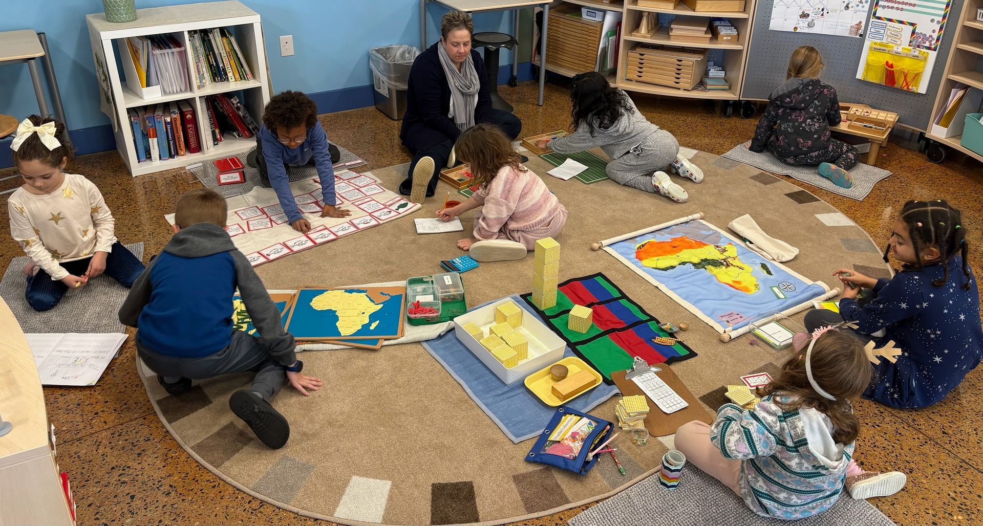 A group of Montessori children and guide are sitting in a circle on the floor working with montessori materials in a classroom.