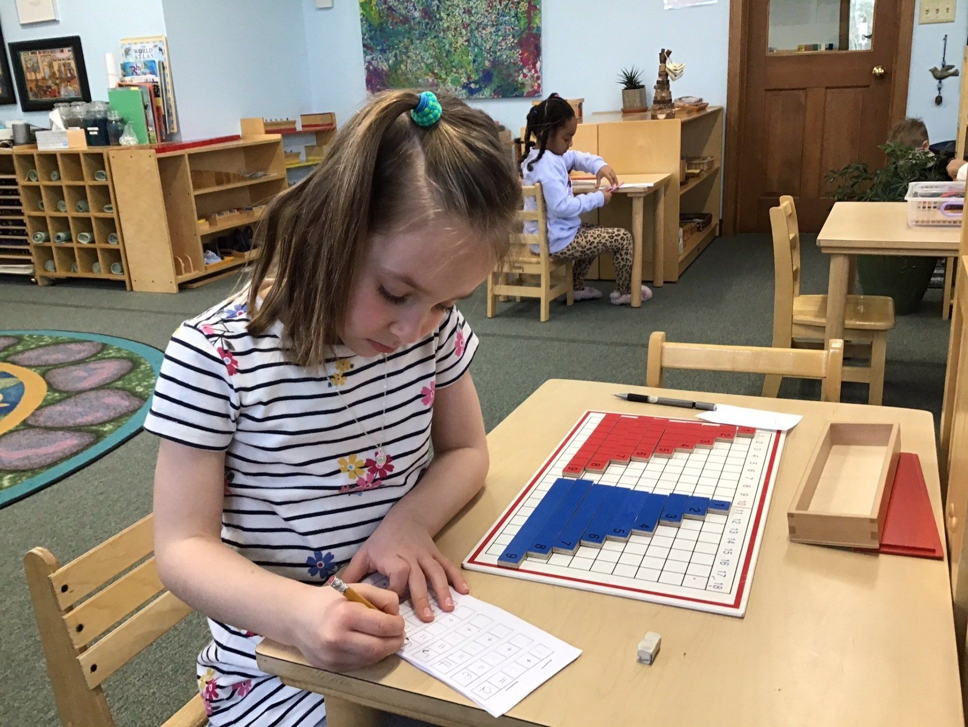 A little girl is sitting at a table writing on a piece of paper with math montessori materials.