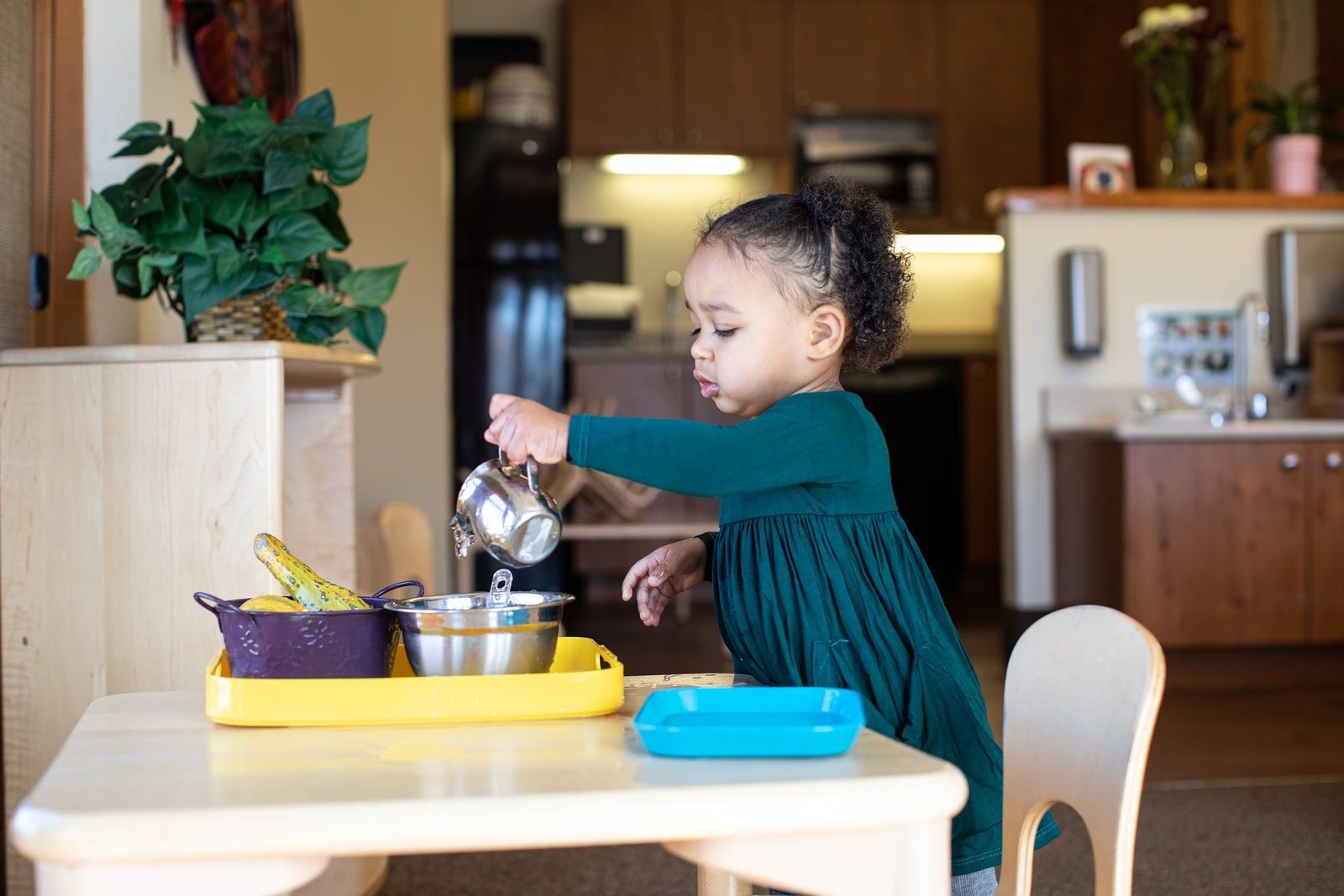 Montessori toddler pouring water from a silver pitcher into a metal bowl at a table.
