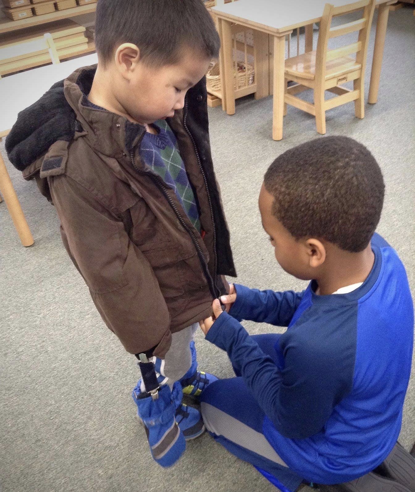 An older boy in a blue shirt helps a younger boy with his jacket fostered by the montessori teaching on kindness and peace.